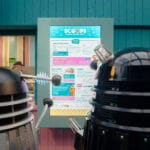 Two Daleks stand in front of an ice cream shop menu board at a sci-fi convention.