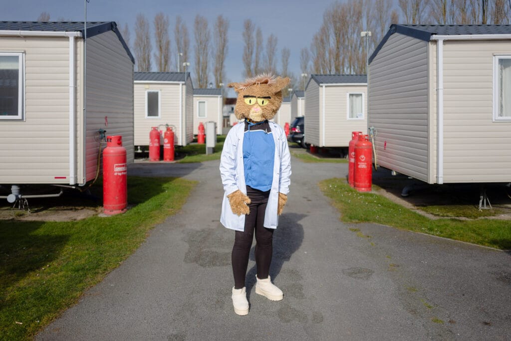 A person wearing a cat costume and a lab coat stands in a parking lot between rows of mobile homes.
