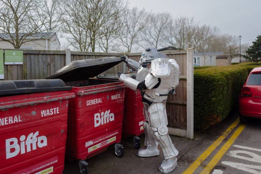 A person in a silver power armor costume is seen opening a red bin labeled "Biffa" at a park. The background shows a wooden fence, some trees and a red car parked on the side.