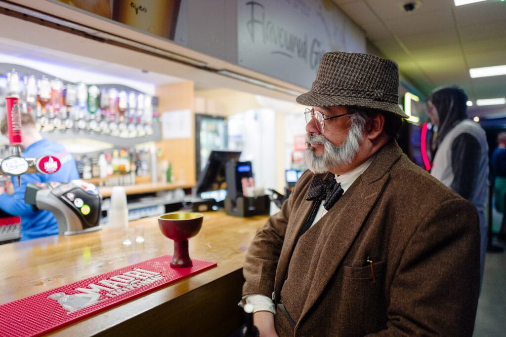A man in a tweed hat and brown suit sits at a bar, looking to the right. He has a gray mustache and goatee. In the background, there is a bar with many bottles and taps, and other people are visible but out of focus.