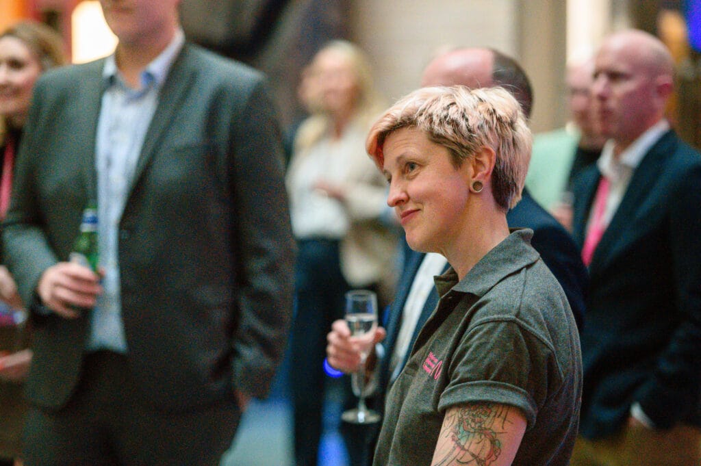 A woman with short pink hair and a tattoo on her arm stands in a room with other people.