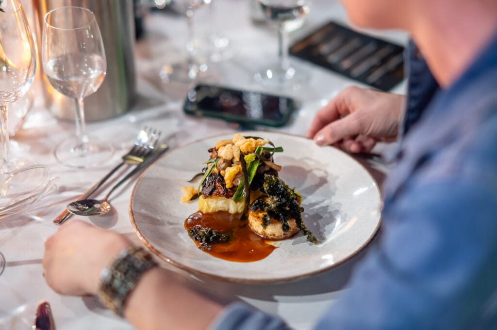 A person is sitting at a table with a plate of food in front of them. The food is a plated dish with meat, potatoes, and vegetables. The person is wearing a blue shirt. There are wine glasses and silverware on the table.