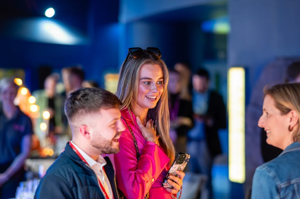 A woman in a pink jacket smiles while looking at her phone. Two other people stand nearby.