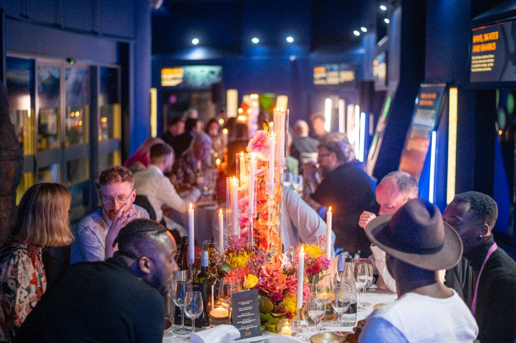 A long table with many people sitting around it in a dark room. There are many candles and flowers on the table.