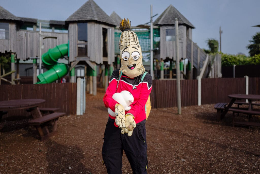 A person in a corn cob costume holds a small white stuffed animal in front of a playground.