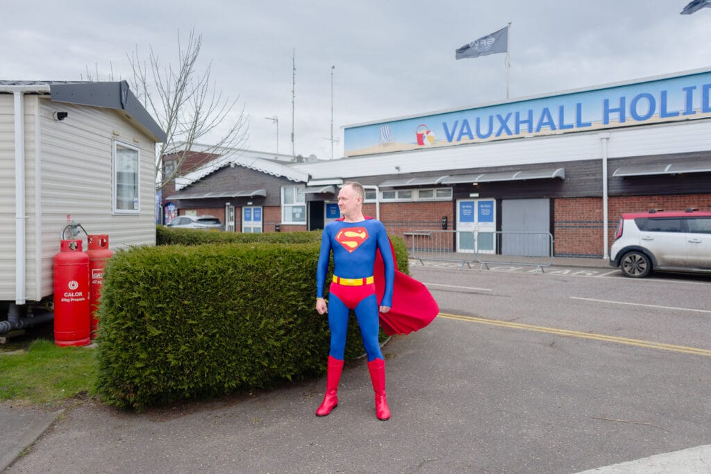 A person dressed as Superman stands in a parking lot outside a building that says "Vauxhall Holiday Park".