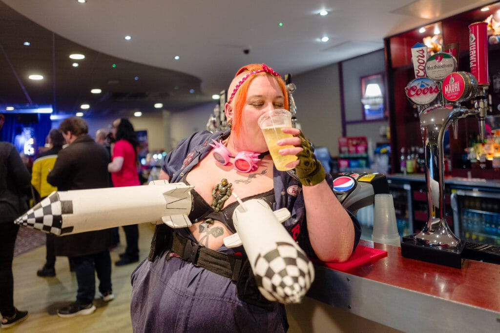 A cosplayer, possibly dressed as Tank Girl, stands at a bar with two rocket-shaped objects strapped to her bra. She is drinking from a plastic cup and smiling slightly. In the background are other people at a convention. The bar has several beer taps visible.