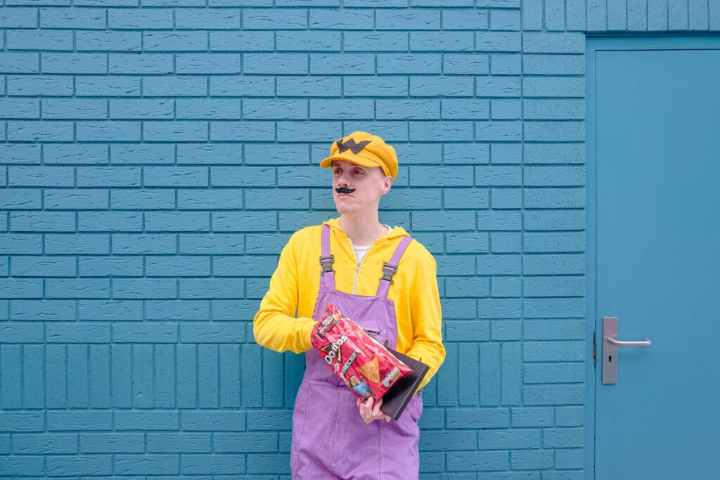 A person dressed in Wario cosplay stands in front of a blue brick wall, holding a bag of Doritos.