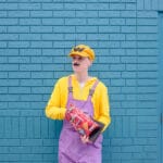 A person dressed in Wario cosplay stands in front of a blue brick wall, holding a bag of Doritos.