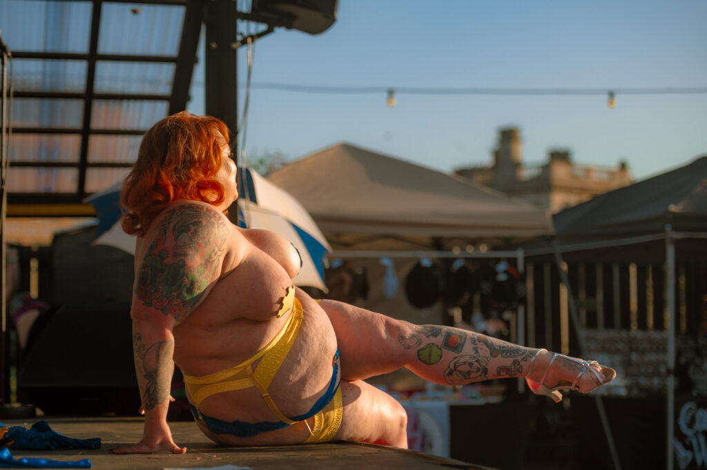 A fat positive performer in a yellow and blue lingerie set, with red hair and tattoos, poses on a stage at Future Yard in Birkenhead.