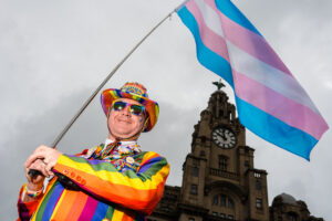 A person in colorful attire holding a transgender pride flag in front of the Liver Building in Liverpool.