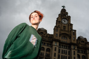 A person with red hair and a nose ring stands in front of the Royal Liver Building in Liverpool, England. The person is wearing a green sweatshirt. The sky is overcast.