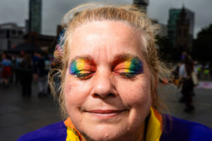 Close-up of a person's face with eyes closed and rainbow-colored eyeshadow, likely at Liverpool Pride. The person is smiling, and the background shows a blurred crowd and buildings.