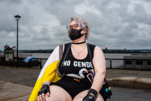 A person wearing a mask and glasses, draped in a non-binary pride flag, sitting outside on a cloudy day. The person is wearing a shirt with the words "NO GENDER" on it.