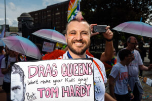 A man with a pink mohawk and a rainbow flag on his face smiles at the camera while holding a sign that says "Drag Queens won't make your kids gay! But Tom Hardy". The image is taken at Liverpool Pride.