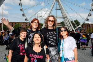 A group of people celebrating Liverpool Pride. The photo shows five smiling individuals in the foreground with a large Ferris wheel in the background. People are wearing pride-themed clothing and accessories, and some are holding flags.