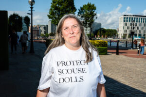 A woman wearing a white t-shirt with the words 'PROTECT SCOUSE DOLLS' printed on it, standing in front of a waterfront with buildings in the background.