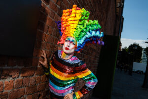 A person in vibrant pride attire, including a rainbow wig and sequined outfit, poses against a brick wall at Liverpool Pride.