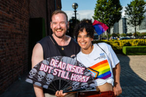 Two people smiling at the camera, holding a pride flag and a fan that says "Dead Inside But Still Horny", at Liverpool Pride.