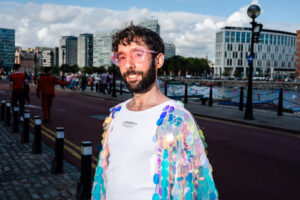 A man with a beard wearing heart-shaped glasses and a sequined jacket smiles at the camera during Liverpool Pride.