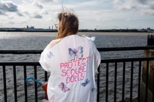 A person stands at the water's edge, wearing a white top adorned with butterfly appliqués and the words 'PROTECT SCOUSE DOLLS' embroidered on the back. The person is facing away from the camera, looking out at the water with a city skyline in the distance. The sky is partly cloudy.