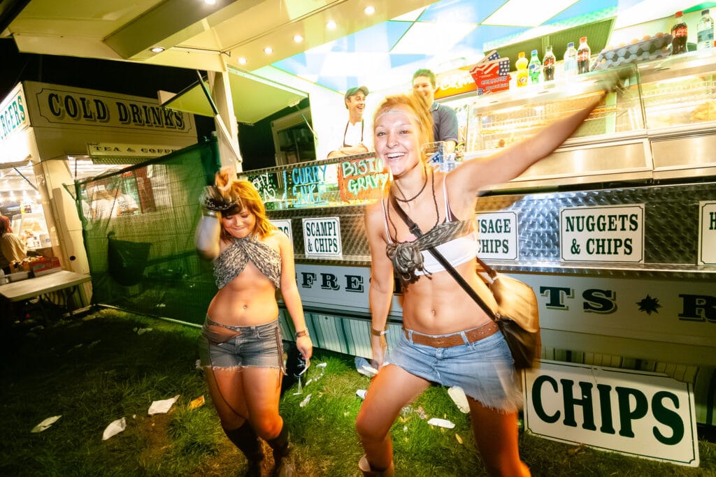 Two young women dancing in front of a food truck at a music festival.