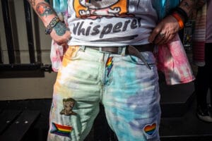 Close-up of a person's torso and arms, adorned with tattoos and colorful accessories, standing at Wirral Pride. They wear a t-shirt with the word 'Whisperer' and jeans decorated with patches including a rainbow heart and a lightning bolt.
