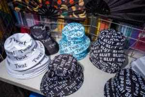 Close-up of a table displaying bucket hats with offensive words printed on them, such as 'Twat' and 'Slut'.