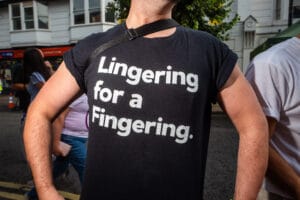 A person wearing a black t-shirt with the words "Lingering for a Fingering" printed on it, standing in a crowd. The photo is taken at Wirral Pride.