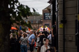 A crowd gathers at Wirral Pride in New Brighton, with people hugging and interacting near a pub with a 'Perch Rock' sign. Two unicorn mascots are visible in the crowd, and the sky is overcast.