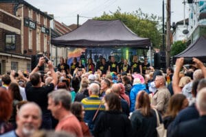 A crowd gathers at Wirral Pride 2025 in New Brighton, with a stage in the background where a choir is performing.