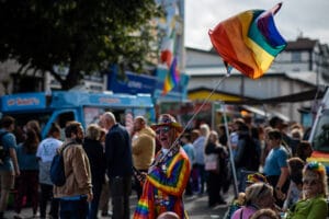 A vibrant photo from Wirral Pride 2025 in New Brighton, featuring a crowd of people, rainbow flags, and a man in colorful attire.