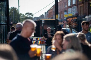 A crowd of people enjoying a sunny day at Wirral Pride in New Brighton. People are holding drinks and flags with rainbow colors are visible.