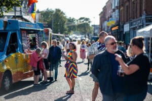 A street scene at Wirral Pride 2025 in New Brighton, featuring a crowd of people, an ice cream van, and rainbow flags.