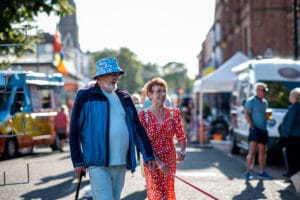 An elderly couple walking down a street during a pride event, the man is wearing a blue jacket and hat, the woman is wearing a red dress with white polka dots.