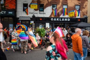 A vibrant street scene from Wirral Pride 2025 in New Brighton, featuring a crowd of people celebrating with rainbow flags and attire, in front of buildings with pride flags and signage.