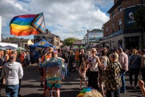 A vibrant street scene at Wirral Pride 2025 in New Brighton, with a large crowd of people, a rainbow flag, and confetti in the air.