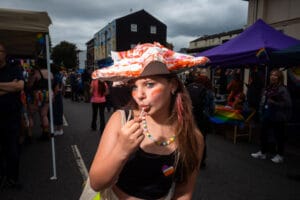 A young person with blue eyes, pink-streaked hair, and a lollipop in their mouth, wearing a creative hat, at Wirral Pride, surrounded by crowds and rainbow flags.