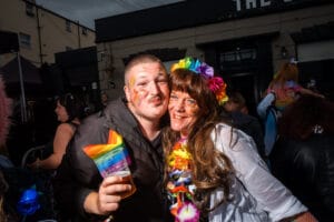 Two people smiling and embracing at Wirral Pride, one holding a rainbow flag cup, with others in the background.
