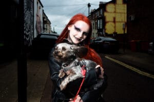A person with bright red hair and dramatic makeup holds a small dog on a leash in the street.