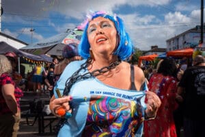 A person with blue hair and makeup is holding up their shirt at a Pride event. The shirt says 'IM LILY SAVAGE THE BLONDE BARS'.