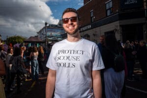 A man wearing sunglasses and a t-shirt that says 'PROTECT SCOUSE DOLLS' smiles at the camera in front of a crowd of people at Wirral Pride.