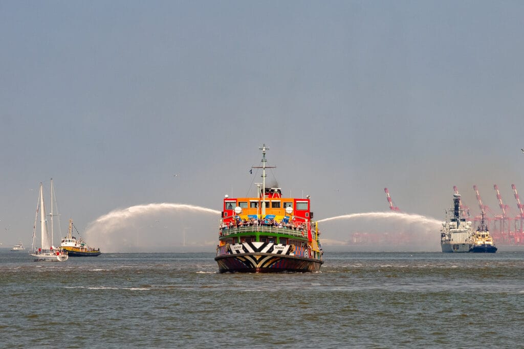 A colourful passenger ferry sails on the Mersey River in Liverpool, with water jets spraying from a fire boat, in front of a backdrop of the city's port and cranes.