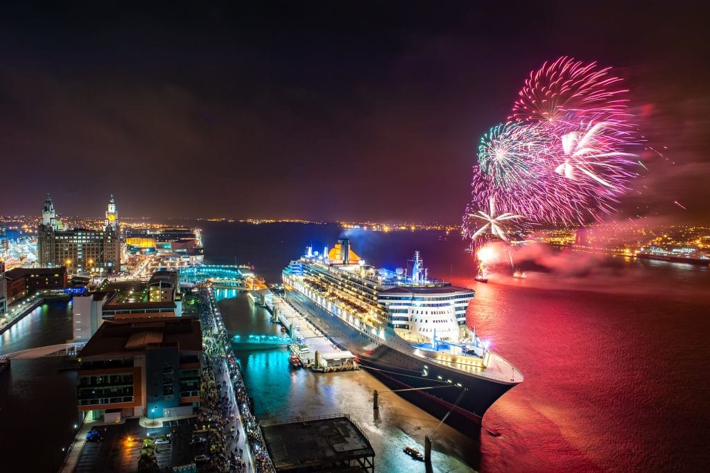 Queen Mary 2 cruise ship docked in Liverpool, England, with fireworks in the night sky.