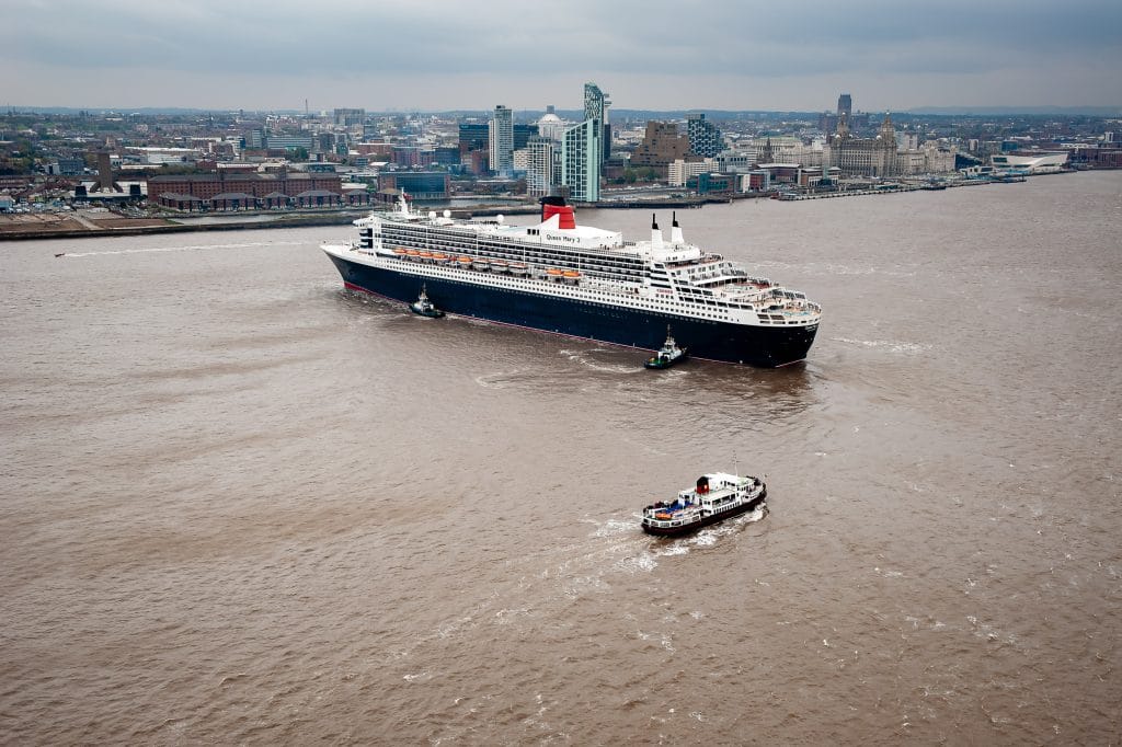 An aerial view of the Cunard cruise ship Queen Mary 2 on the River Mersey, with the Liverpool skyline in the background.