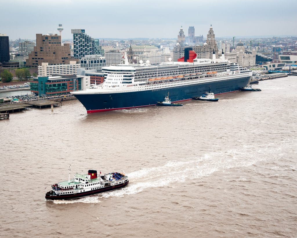 Aerial view of the Cunard cruise ship Queen Mary 2 arriving in Liverpool, England, with tugboats assisting and a ferry boat in the foreground.
