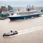 Aerial view of the Cunard cruise ship Queen Mary 2 arriving in Liverpool, England, with tugboats assisting and a ferry boat in the foreground.