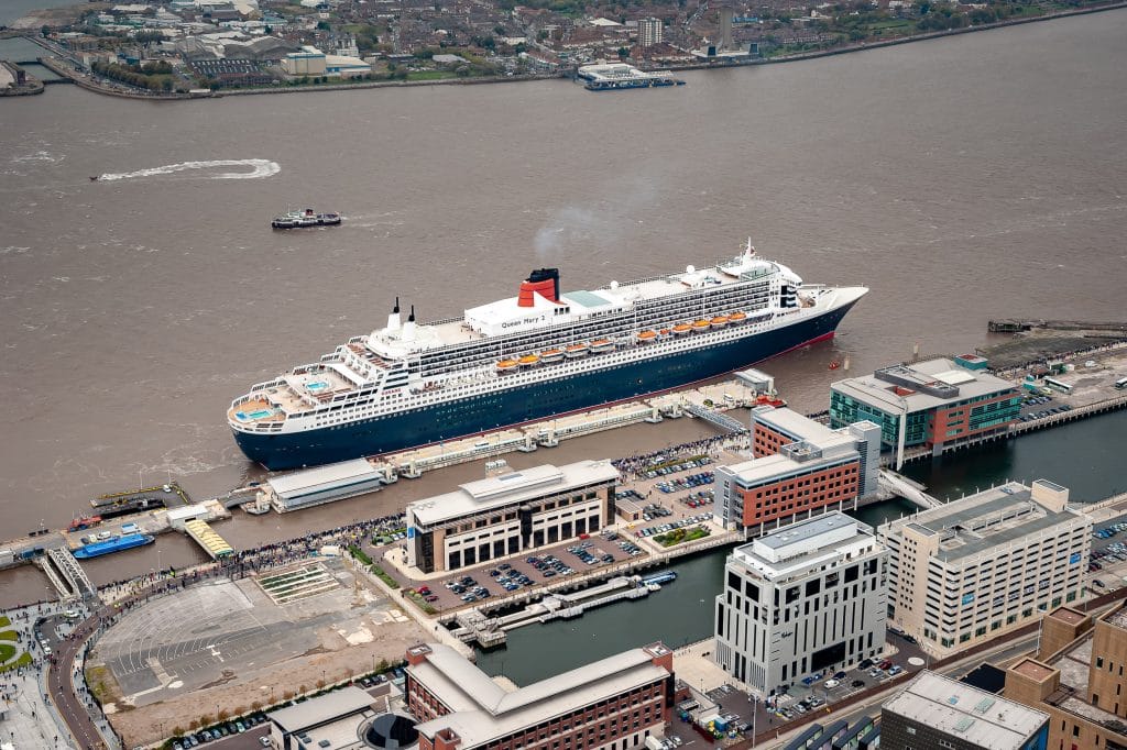 Aerial view of the Cunard cruise ship Queen Mary 2 docked at Liverpool Pier Head, with surrounding buildings and crowds of people.