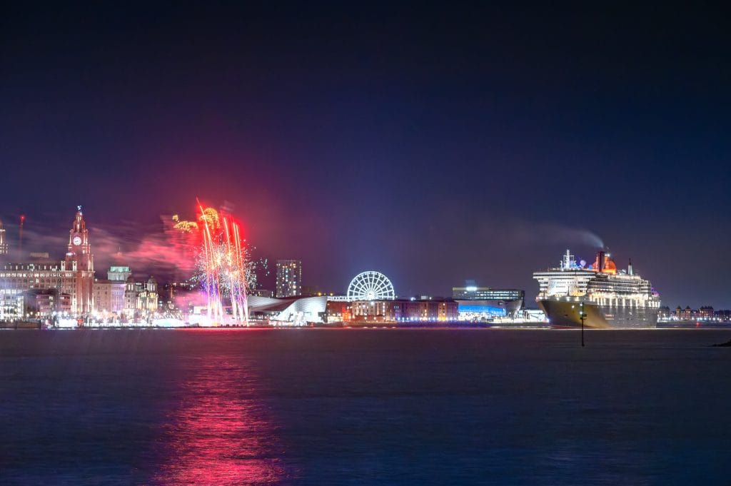 Night view of the Cunard cruise liner QM2 leaving Liverpool, with fireworks and city skyline in the background.