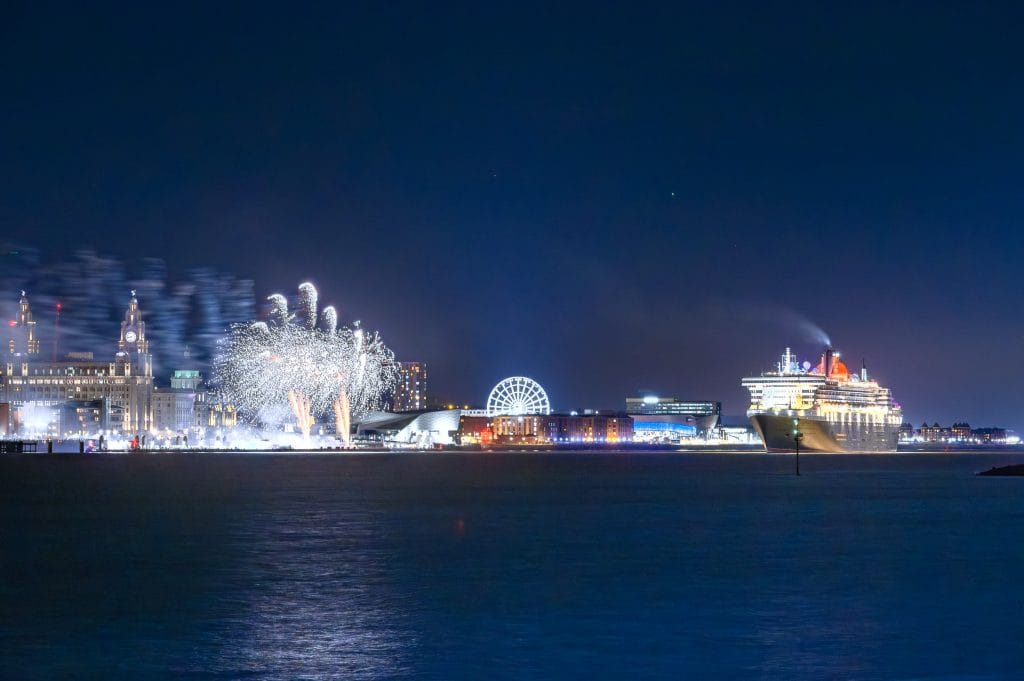 The Cunard cruise liner QM2 sails away from Liverpool at night, with fireworks in the sky and illuminated buildings along the waterfront.